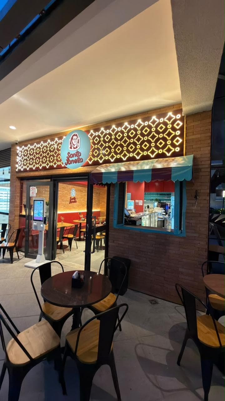 A fachada da unidade café à noite: o painel iluminado com o símbolo da Santa Janela, a porta aberta e a janela de atendimento com toldo listrado.